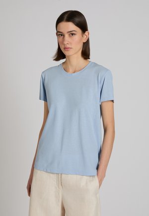 Frau mit glattem, dunklem Haar trägt hellblaues kurzärmeliges Hemd und cremefarbene Hose, steht vor einem schlichten hellgrauen Hintergrund.