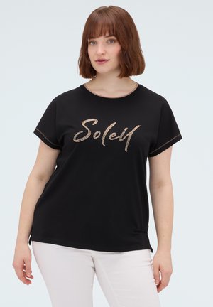 Kvinde med rødt hår iført sort T-shirt med teksten "Soleil" i guldskrift og hvide bukser, stående mod en lys baggrund.
