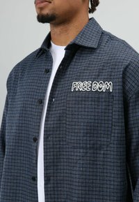 Blauw en grijs geruit overhemd met een knoopsluiting, voorzien van een borstzak en witte geborduurde tekst "FREE DOM." Zachte stoftextuur.