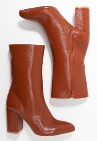 Une paire de bottes mi-mollet brillantes en orange brûlé avec des talons carrés et des fermetures éclair sur le côté, posées à plat sur un fond blanc.