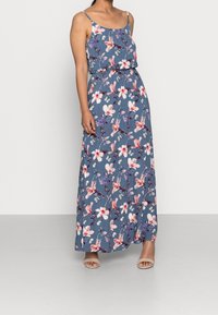 Kvinna som bär en ärmlös blå maxiklänning med rosa och vita blommönster och beige öppen-tå klackar, stående mot en enfärgad bakgrund.