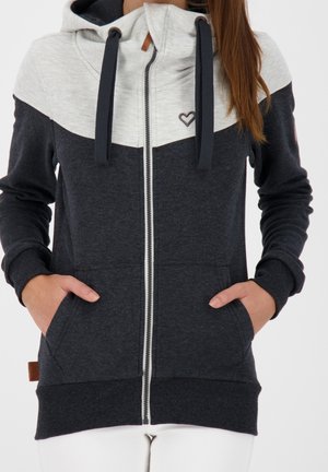 Kobieta ubrana w dwukolorową czarno-szarą bluzę z kapturem na zamek, z emblematem serca i rękami w przednich kieszeniach.