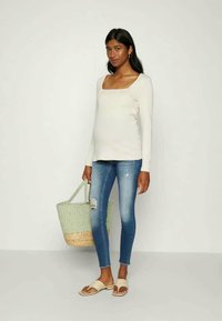 Blusa de mangas longas bege clara com decote quadrado, jeans skinnies azuis com detalhes desgastados, bolsa de palha verde e sandálias bege.