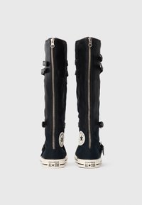 Hohe schwarze Converse All Star Stiefel mit Rückenreißverschlüssen, Knöchelriemen und weißen Gummisohlen, von hinten auf einem weißen Hintergrund gezeigt.