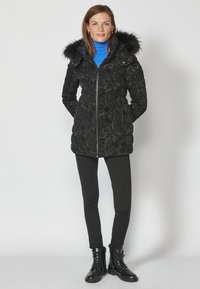 Melna quiltota parka ar kažokādas oderi kapuci, ar rāvējslēdzēju un dekoratīvu paisley rakstu. Valkāta virs zilā turtleneck un melnajām legingām.