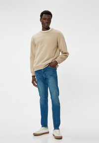 Sudadera beige con cuello redondo, jeans azul marino y zapatillas blancas con suelas marrones. El modelo está de pie frente a un fondo blanco liso.