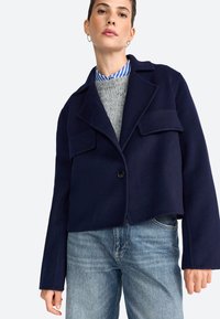 Giacca tagliata navy in lana con collo a scialle, due tasche frontali, chiusura con un bottone e maniche ampie sopra un maglione grigio.