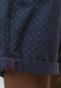 Pantalones cortos azul marino con un sutil patrón de diamantes, que presentan un dobladillo enrollado y un acento de costura en rosa en el puño. Fabricados con un tejido ligero.
