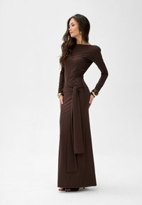 Brązowa sukienka z długim rękawem wykonana z elastycznego materiału, z dopasowanym krojem, szczegółem w postaci węzła po bokach w pasie oraz długością do podłogi.