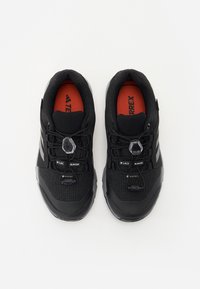 Black outdoor shoes featuring a textured fabric upper, toggle lacing system, gray accents, and a red insole with logo detail.
