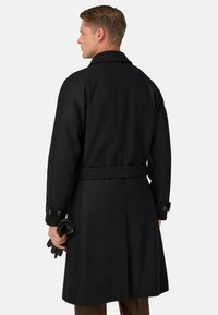 Vīrietis redzams no mugurpuses, ģērbies melnajā jostotajā mētelī un kreisajā rokā turot melnas ādas cimdus uz vienkārša fona.