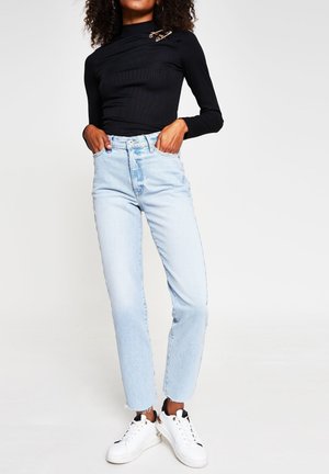 Femme debout les mains dans les poches, portant un haut noir à manches longues, un jean taille haute bleu clair et des baskets blanches sur fond blanc.