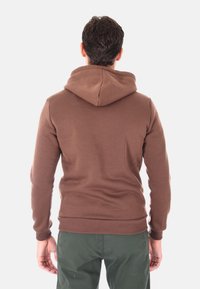 Uomo in piedi con le spalle alla fotocamera, indossa un felpa marrone e pantaloni verdi scuri su uno sfondo bianco semplice.