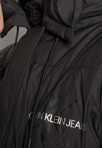 Svart puffjacka med hög krage, strukturerad tyg och reflekterande "CALVIN KLEIN JEANS"-logotyp på framsidan. Har en dragkedja och snörning.