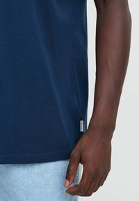 T-maica mornarice iz bombaža z zrnasto površino, s stranskim razporkom in majhnim označevalnim logotipom na robu, v kombinaciji z svetlim denimom.