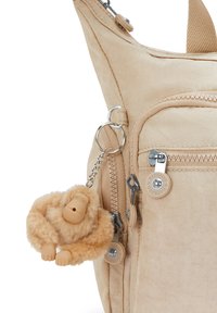 Zaino in tessuto beige con tasca frontale con cerniera e un piccolo portachiavi di una scimmietta di peluche beige attaccato a un anello metallico.