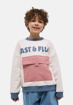 Enfant souriant aux cheveux bouclés portant un sweat-shirt beige et bleu avec le texte "FAST & FLU" et une grande poche rose, accompagné d'un jean bleu ample.