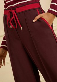 Burgundské široké kalhoty s červeným šňůrkovým pasem, obsahující boční kapsy a kontrastní červené detaily. Hladká textura a uvolněný střih.