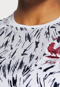 Camiseta deportiva blanca con un patrón abstracto en negro y gris, cuello redondo y un logotipo de gallo bordado en rojo en el pecho.