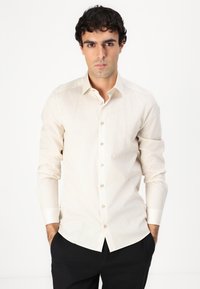 Cămașă bej cu mânecă lungă, cu un model texturat subtil, cu închidere buton și guler standard. Purtată cu pantaloni negri, mâinile în buzunare.