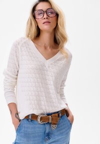 Witte gebreide trui met een gemêleerd hexagonaal patroon en een V-halslijn. Gepaard met blauwe jeans en een bruine riem met een decoratieve gesp.