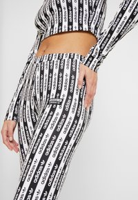 Svart och vit atletisk set som består av en kortärmad topp med lång ärm och högmidjade leggings, båda med ett upprepande "adidas"-logotyp och trefoil-mönster.