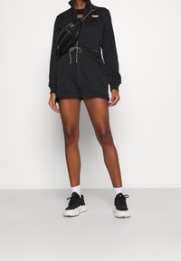 Černý overal na zip s dlouhými rukávy, stahovacími šňůrkami v pase a kulatým výstřihem. Nosený s černými teniskami a bílými ponožkami. Minimalistický design.