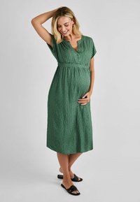 Groene zwangerschapsjurk met knoopsluiting aan de voorkant, korte mouwen en een gathers op de taille. Heeft een gestructureerde stof met een klein stippenpatroon. Zwarte sandalen.