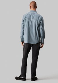 Camisa de manga larga a rayas azul claro con borde curvado, puños abotonados, combinada con jeans oscuros y zapatos negros, vista desde atrás.