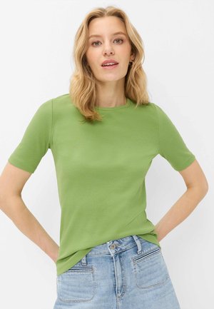 Blonde Frau mit welligem Haar trägt grünes Kurzarm-Top und hellblaue Jeans, steht mit den Händen in der Hüften vor weißem Hintergrund.