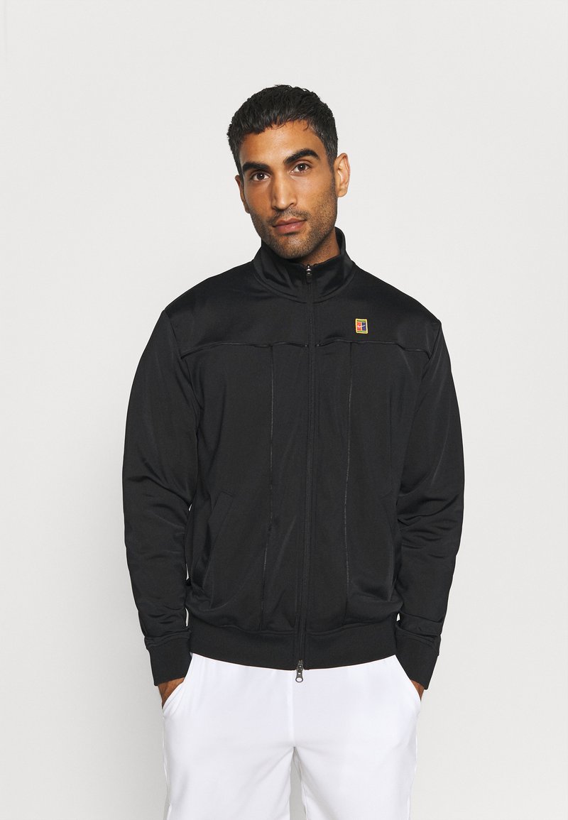 Chaqueta negra con cremallera y cuello alto, que cuenta con dos bolsillos laterales y un pequeño logotipo multicolor en el lado izquierdo del pecho. Textura de tela suave.