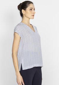 Biała bluzka z niebieskim wzorem, z krótkim rękawem i dekoltem w kształcie V. Tkanina jest gładka, z bocznymi rozcięciami dla swobody ruchów.