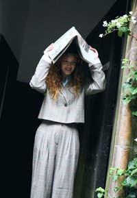 Femme en costume à carreaux gris souriante, tenant un journal ouvert au-dessus de sa tête près d'une porte sombre avec des plantes vertes sur le côté.