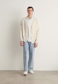 Krémový pletený kardigan s knoflíky, oblečený přes bílou košili. Světle modré džíny a bílé tenisky dokončují outfit.