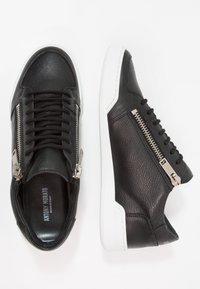 Černé kožené tenisky s bílými gumovými podrážkami, vybavené bočními zipy, texturovaným svrškem a černými tkaničkami. Minimalistický design s výraznými prvky.
