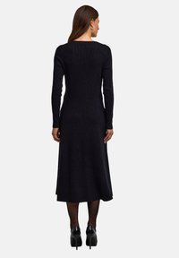 Abito in maglia nero a maniche lunghe con corpetto aderente e gonna svasata, caratterizzato da uno scollo a schiena scoperta e una texture morbida, abbinato a scarpe con tacco alto.