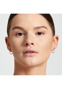 Donna con pelle chiara e trucco leggero, che indossa piccoli orecchini a cerchio d'oro e un orecchino a perno, con capelli lisci e sfondo neutro.
