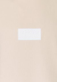 Beige fabric with a centered white rectangular Adidas logo patch featuring the brand name and trefoil symbol twice.