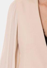Blazer beige con maniche morbide efluenti, caratterizzato da un design strutturato e una texture liscia, che mette in mostra uno stile minimalista.