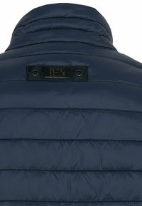 Veste matelassée bleu marine, vue de dos, avec un col montant et une étiquette noire rectangulaire cousue près de l'encolure.
