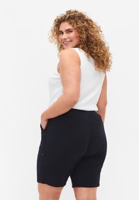 Melni šorti ar elastīgu jostasvietu, sānu kabatām un gludu tekstūru, apvienoti ar balto bez piedurknēm topu. Modelis smaidot.