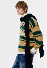 Chlpatý béžový sveter so zelenými pruhmi, s výstrihom do V a logom, doplnený o textúrovaný čierny šál, nosený cez bielu košeľu a džíny.