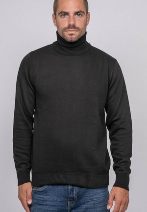 Homme avec une barbe courte portant un pull à col roulé noir et un jean bleu, debout devant un fond uni gris clair.