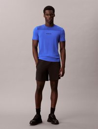 T-shirt atlético azul com mangas curtas e um painel superior em rede, combinado com calções pretos e sapatilhas pretas, usado por um modelo em pé.