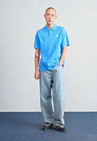 Modrá polokošeľa, svetlo umývané džínsy, čierne tenisky. Polokošeľa má golier a malé logo; džínsy majú voľný strih. Jednoduché farby, minimálne detaily.