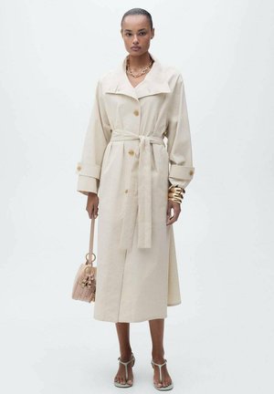 Beige trench coat with large collar and front buttons, waist tie, long sleeves, and a flowing design. Paired with a pink handbag and sandals.