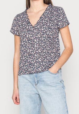 Marineblauwe bloes met korte mouwen, voorzien van een geschulpte V-halslijn, roze en witte bloemmotief, en lichtgewicht stof. Gecombineerd met lichte jeans.