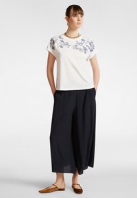 Haut blanc à manches courtes avec broderie florale bleue, associé à un pantalon large noir et des sandales plates marron. Design simple et coupe décontractée.