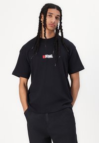 Black cotton t-shirt featuring a central white and red logo. Short sleeves, relaxed fit, and soft texture, paired with dark pants.
