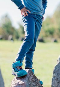 Blå sportsbukser og en matchende sweater, med turkise vandrestøvler der har sorte snørebånd og orange detaljer, stående på en sten.